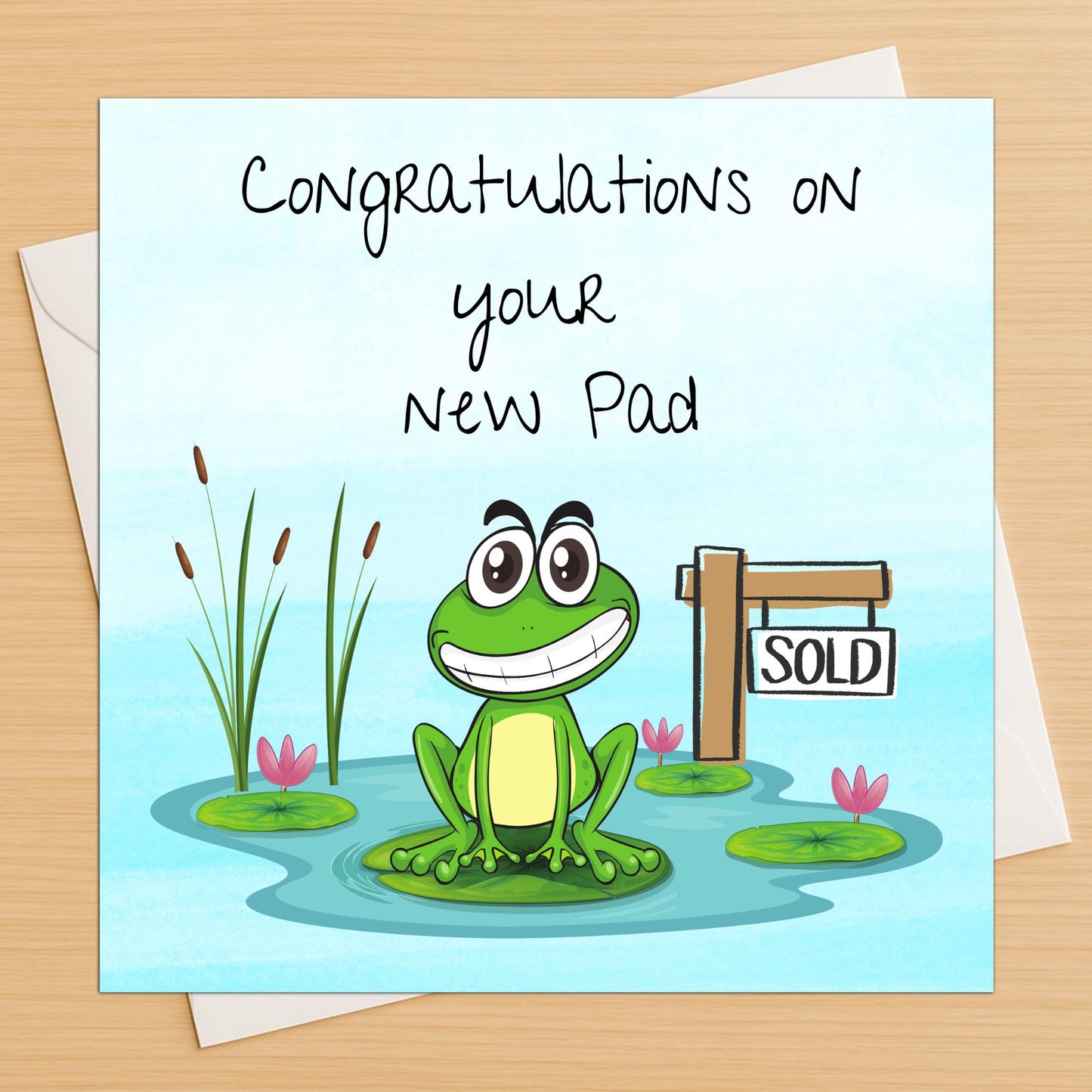 Card with a cartoon frog and 'Congratulations on your new pad' text on a wooden surface